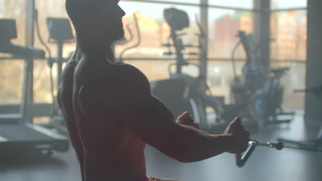 Athlete-doing-exercises-in-the-simulator-with-weights-and-pulls-the-bar-sitting-on-the-background-of-a-large-window-GYM.-Strong-purposeful-athlete-view-from-the-back